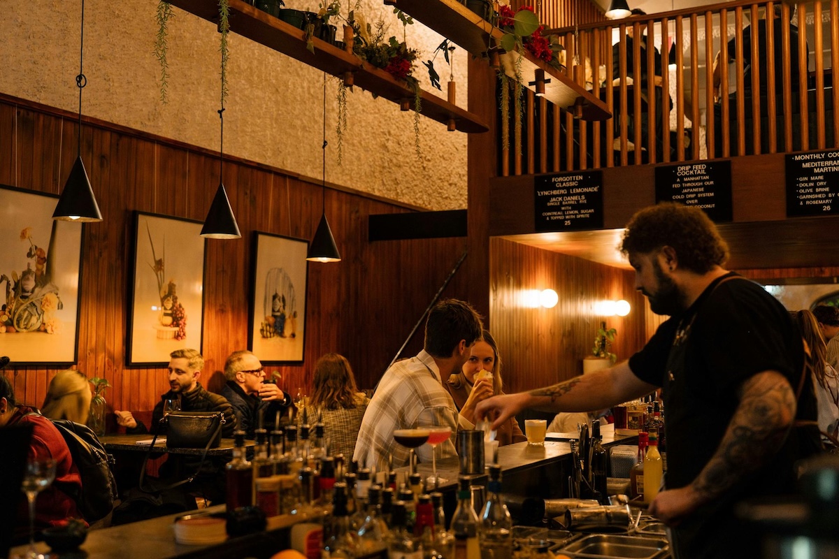 Warmly lit interior of Malt & Juniper bar with patrons seated at tables and a bartender preparing drinks at the counter.